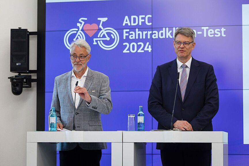 ADFC Fahrradklima-Test 2024 - Frank Masurat und Patrick Schnieder Zwei Männer stehen an Rednerpulten vor einer Präsentationsfläche mit dem Logo des ADFC Fahrradklima-Tests 2024.